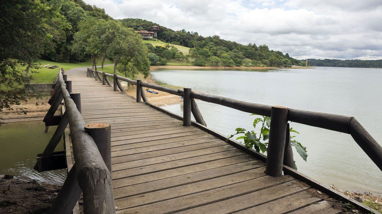 Passarela de madeira com corrimão de troncos à beira do lago do Parque Passaúna, em Curitiba, com morros cobertos de mata ao fundo