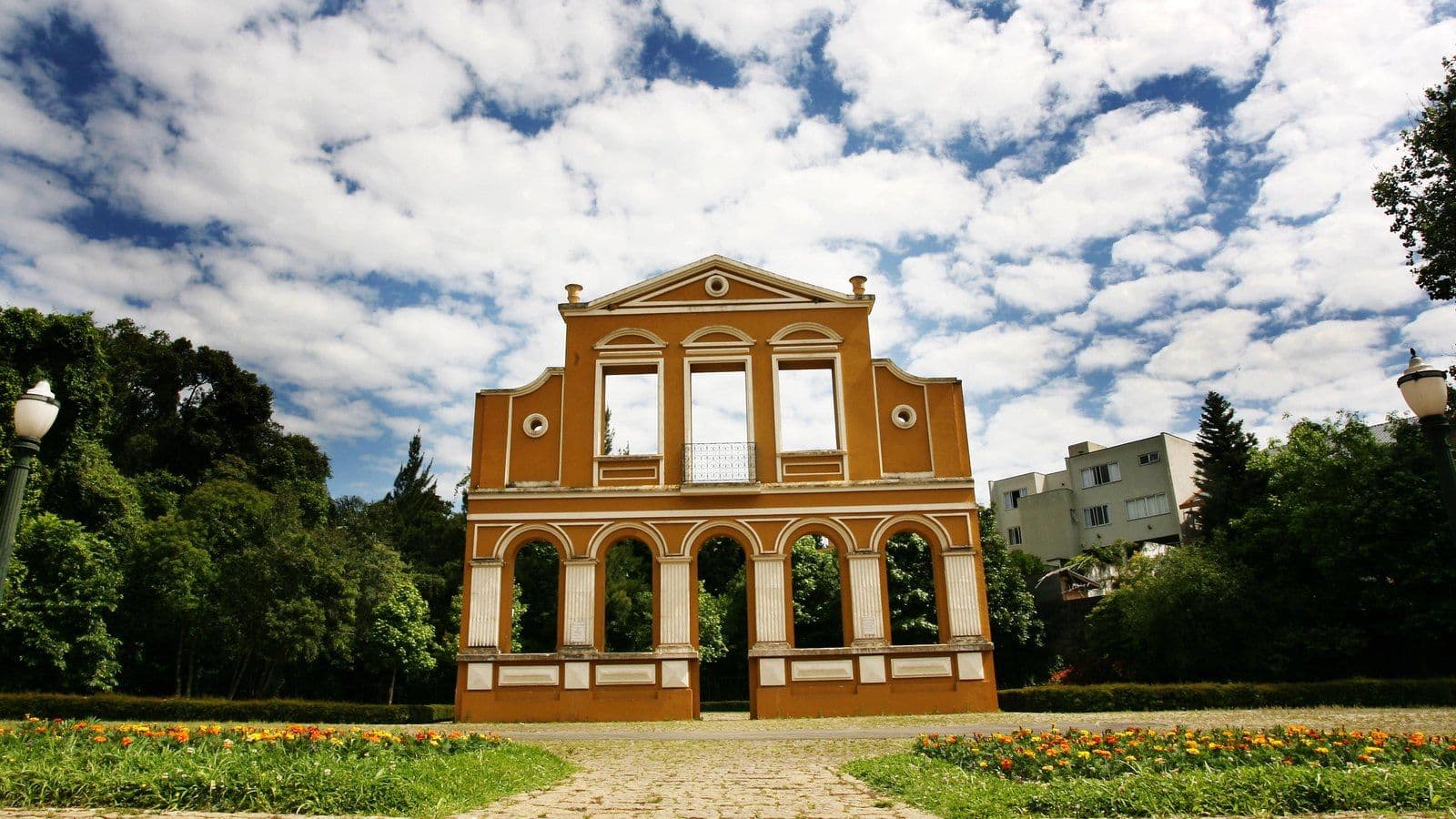 Fachada amarela com arcos e janelas no Bosque Alemão, em Curitiba, entre canteiros de flores, árvores e céu de nuvens