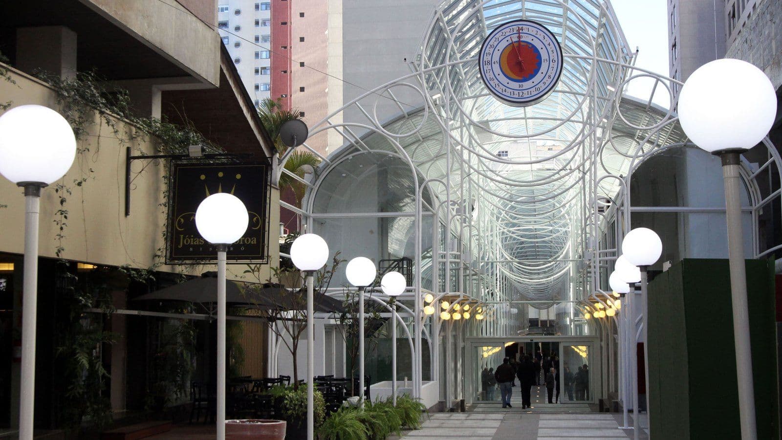 Entrada da Rua 24 Horas, em Curitiba, com arcos de metal branco, cobertura de vidro, relógio de 24 horas e postes com globos de luz