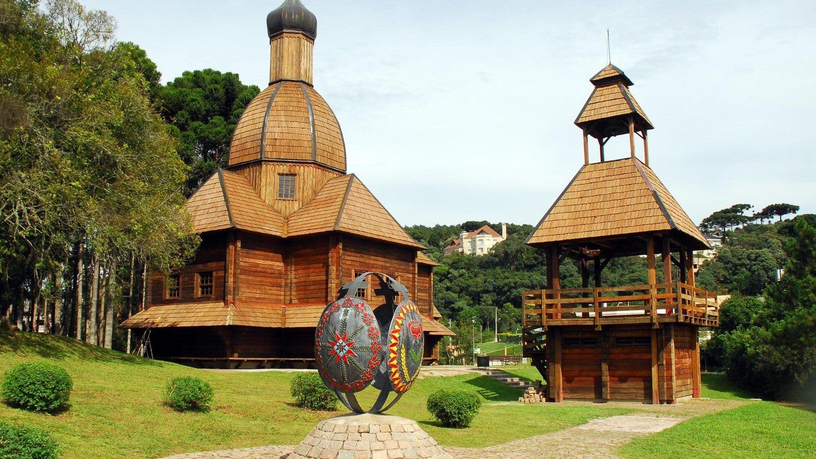 Igreja de madeira com cúpula e campanário do Memorial Ucraniano, no Parque Tingui, em Curitiba, com a escultura de uma pêssanka no gramado